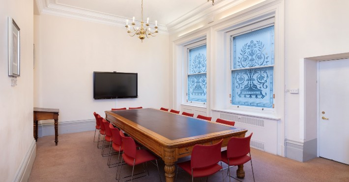 white room with two sash windows, a large table with red chairs around it and a TV screen on the wall
